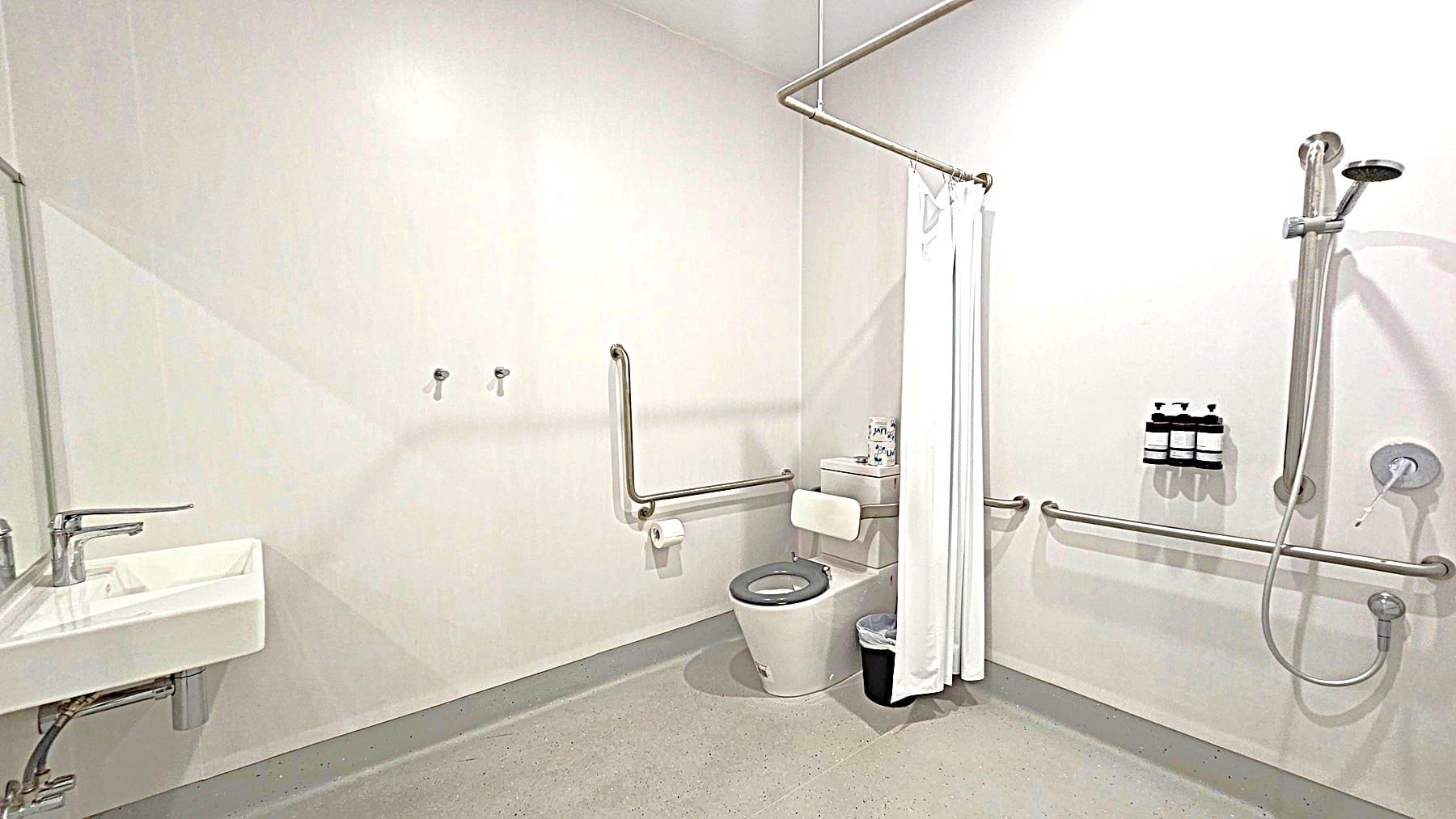A bathroom with a white sink and a metal tap on the left. In the centre, a toilet with a support rail and a roll of toilet paper. A white curtain hangs nearby. On the right, a shower area with a handheld showerhead and three bottles mounted on the wall. The floor is light grey, and the walls are plain white. Metal support bars are installed throughout the room.