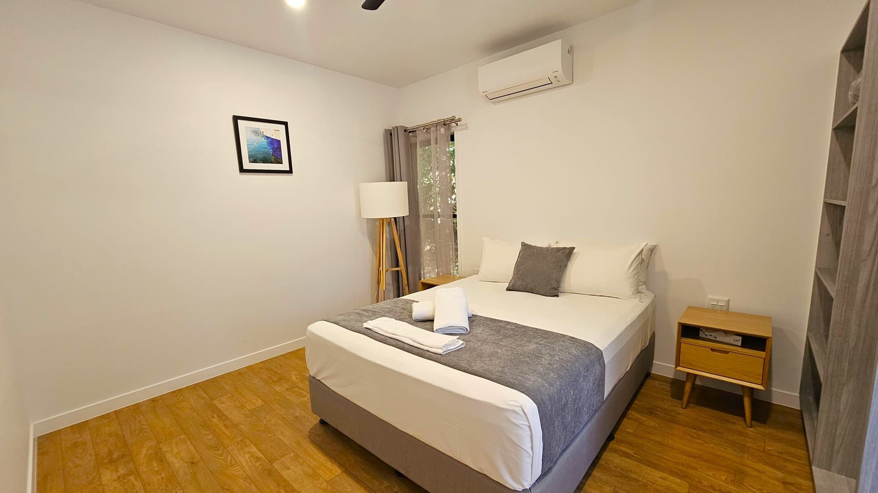 A small bedroom with wooden flooring. On the right, a bed with white and grey bedding, topped with neatly folded towels. A wooden bedside table with a drawer is next to the bed. Above, a wall-mounted air conditioning unit. On the left, a floor lamp with a tripod base stands near a window with sheer curtains. A framed picture hangs on the wall. In the background, a built-in shelf is partially visible. The ceiling is plain with a light fixture.