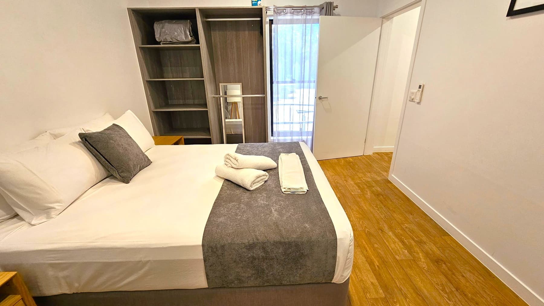 A bedroom with a neatly made bed on the left, featuring white sheets, a grey throw, and two rolled towels. A grey cushion is placed on the bed. To the right, there is a wooden floor and a partially open door leading to a balcony with sheer curtains. In the background, an open wardrobe with shelves contains a folded blanket and some books. The walls are plain white, and a small control panel is visible near the door.