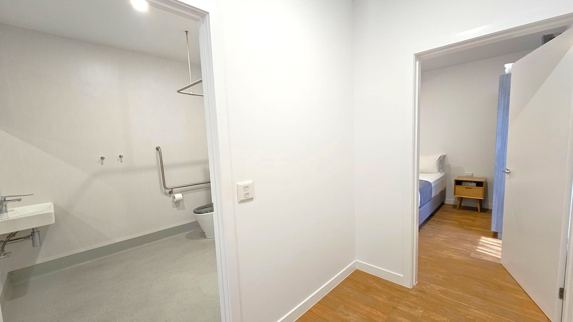 A hallway with two open doors. On the left, a bathroom with a light-coloured floor, a wall-mounted sink, and a toilet with a grab bar. On the right, a bedroom with a wooden floor, a bed with a blue and white bedspread, and a small wooden bedside table. The walls are white, and the ceiling has a recessed light.