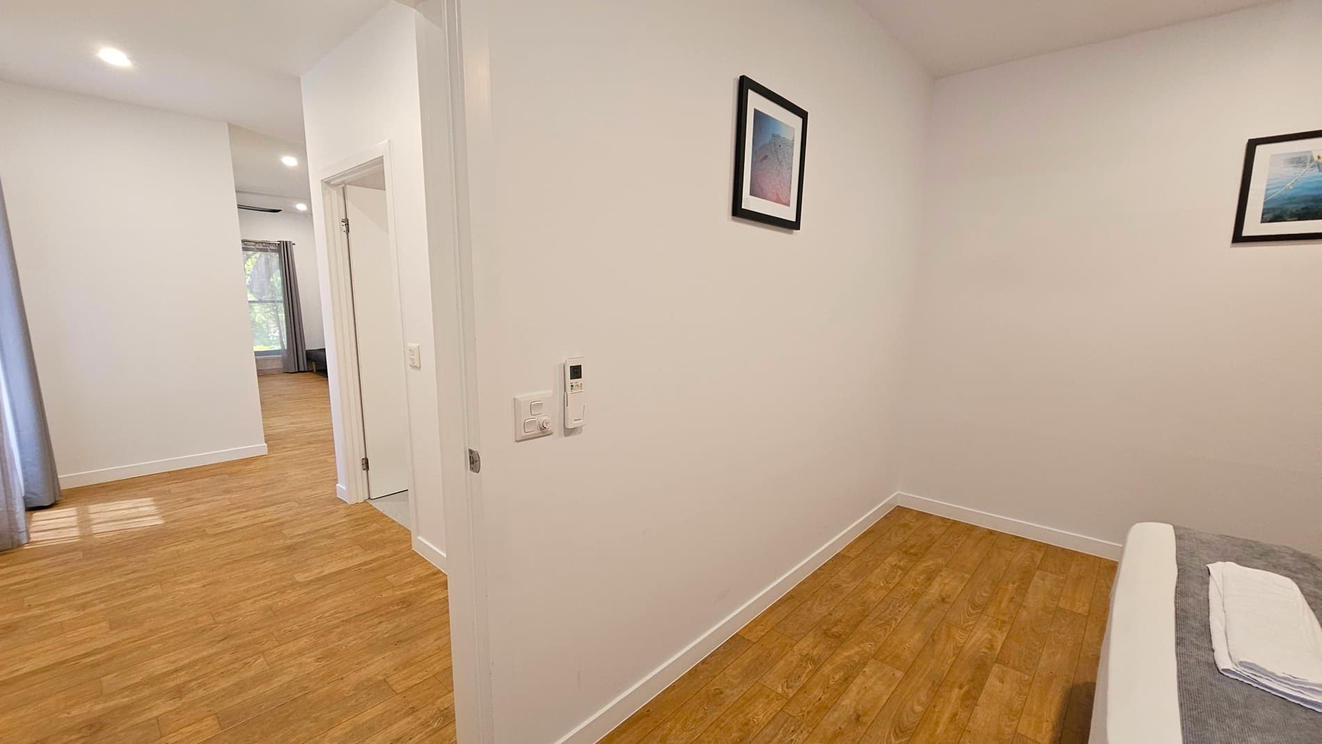 A hallway with light wooden flooring and white walls. On the right, a small section of a bed with a folded towel is visible. A framed picture hangs on the wall above the bed. On the left, the hallway leads to another room with a large window covered by curtains. The ceiling has recessed lighting, and a thermostat is mounted on the wall near the doorway.