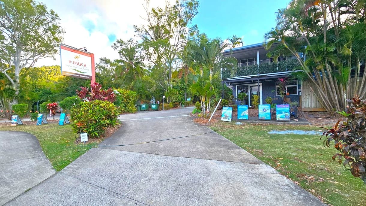 A driveway leads to a building on the right, partially obscured by lush greenery and palm trees. Several signs and posters are displayed along the path. On the left, a large sign reads "Kipara Tropical Rainforest Retreat" with contact information. The area is surrounded by dense foliage, including trees and shrubs. The ground is a mix of concrete and grass, with a small "Exit Only" sign visible. The sky is partly cloudy.