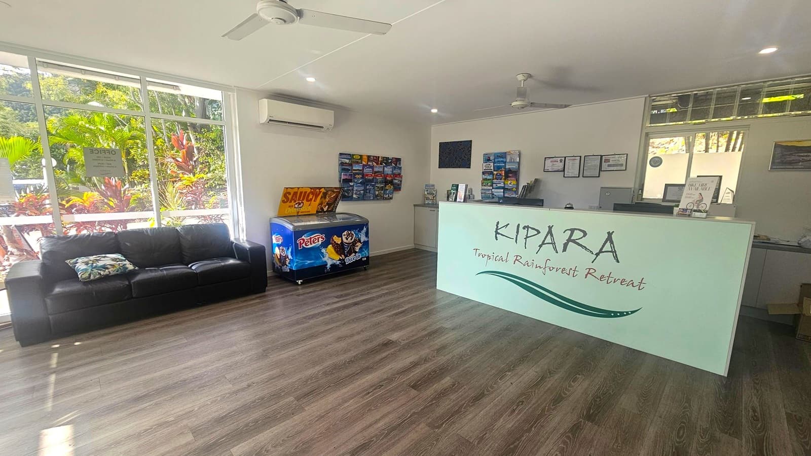 Reception area with a large window on the left, showing lush greenery outside. A black leather sofa with a patterned cushion is positioned in front of the window. To the right, a white reception desk with "KIPARA Tropical Rainforest Retreat" written on it. Behind the desk are brochures and framed certificates on the wall. A ceiling fan and air conditioning unit are visible above. The floor is wooden, and the ceiling is white with recessed lighting.