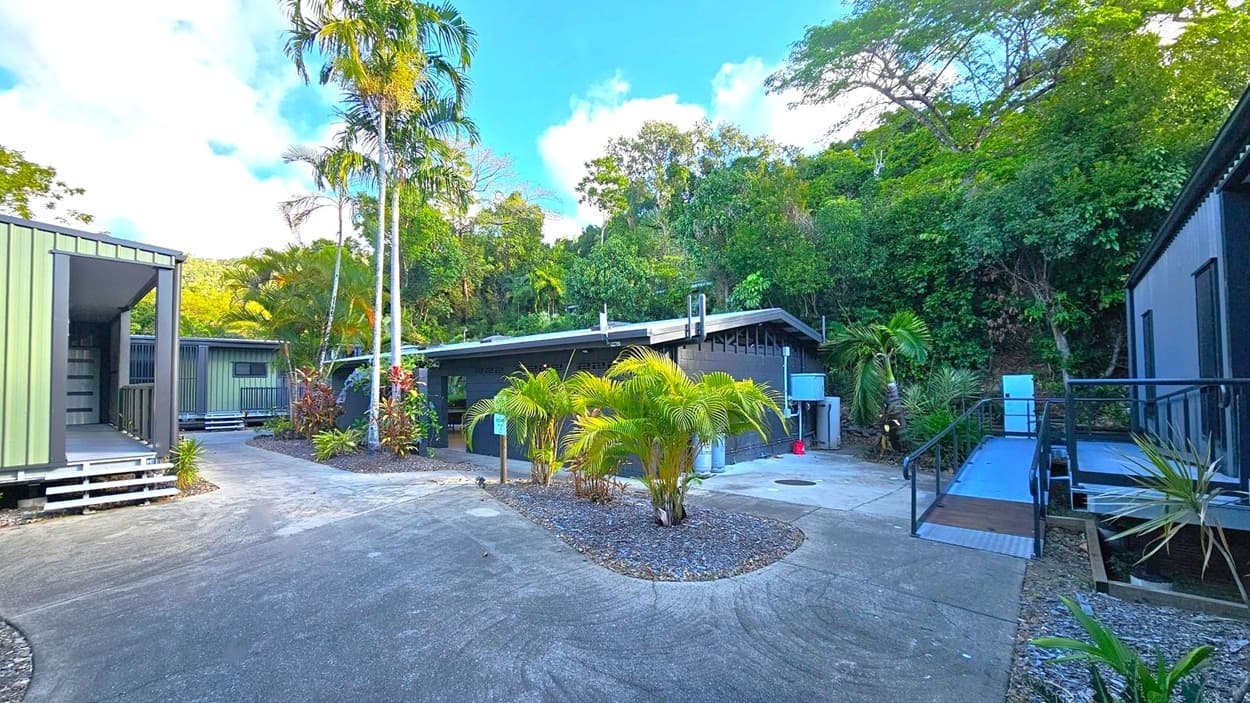 A courtyard with a concrete path surrounded by lush greenery. In the centre, there are small palm trees and plants in a landscaped area. On the left, a green building with a small porch and steps. In the middle, a low, dark building with a sloped roof. On the right, another building with a ramp leading to the entrance. The background is filled with dense trees and foliage.
