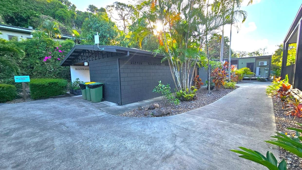 A small building with dark walls is surrounded by lush greenery and palm trees. On the left, a sign points towards the laundry and BBQ area. Two bins are placed near the building. A concrete path curves around the building, leading to another structure in the background. The area is landscaped with various plants and shrubs, and sunlight filters through the trees, creating a serene atmosphere.