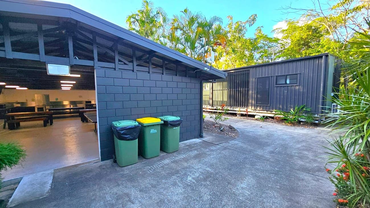 On the left, a building with a dark brick wall and an open entrance reveals a room with tables and bright ceiling lights. Three wheelie bins with green and yellow lids are placed against the wall. On the right, a black corrugated metal structure is elevated on a platform, with small plants around its base. The background features tall palm trees and dense greenery. The ground is paved with concrete, and there are small shrubs along the edges.