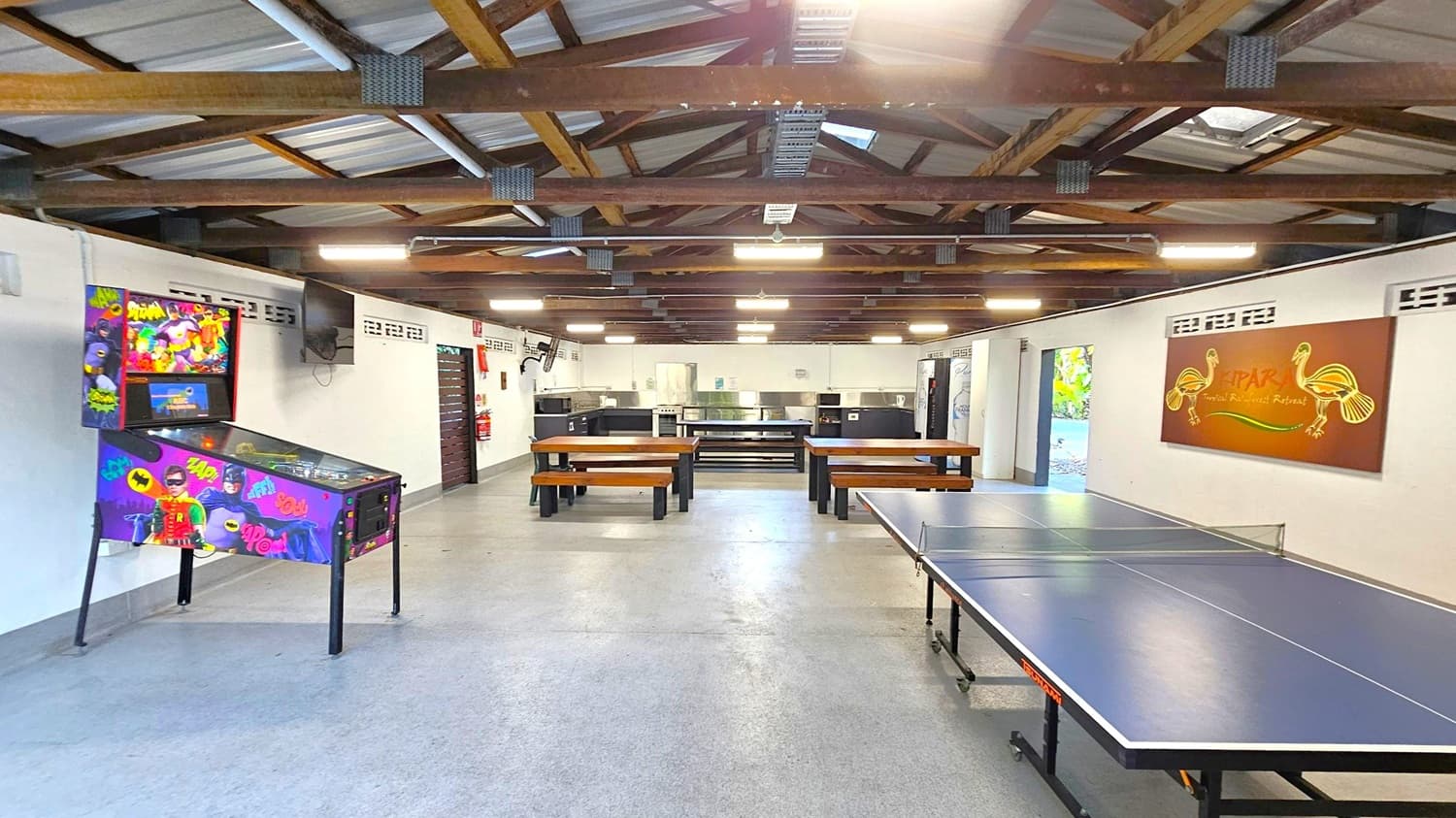 A recreational room with a pinball machine on the left featuring colourful superhero graphics. In the centre, there are wooden picnic-style tables and benches. The right side has a table tennis setup. The back wall has a kitchen area with stainless steel appliances and a large sign with artistic designs. The ceiling is open with exposed wooden beams and fluorescent lighting. The floor is grey and smooth.