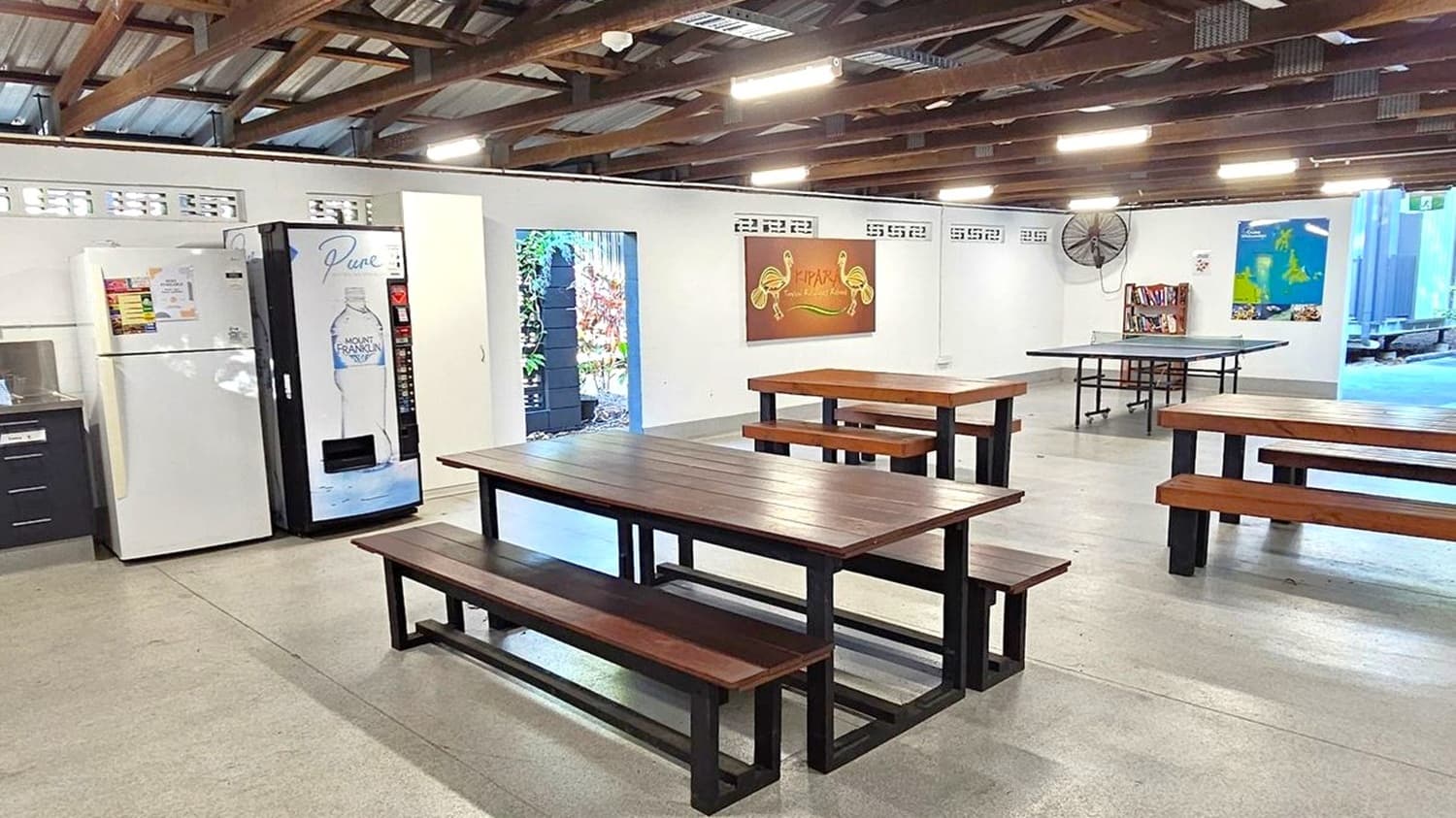 An open-air dining area with a high, exposed beam ceiling. On the left, there's a kitchen counter with a sink, a white refrigerator, and a vending machine. In the centre, wooden tables with benches are arranged on a concrete floor. The right side features a wall with colourful artwork and a fan. The background shows an open entrance leading to an outdoor area with greenery and a vibrant mural.