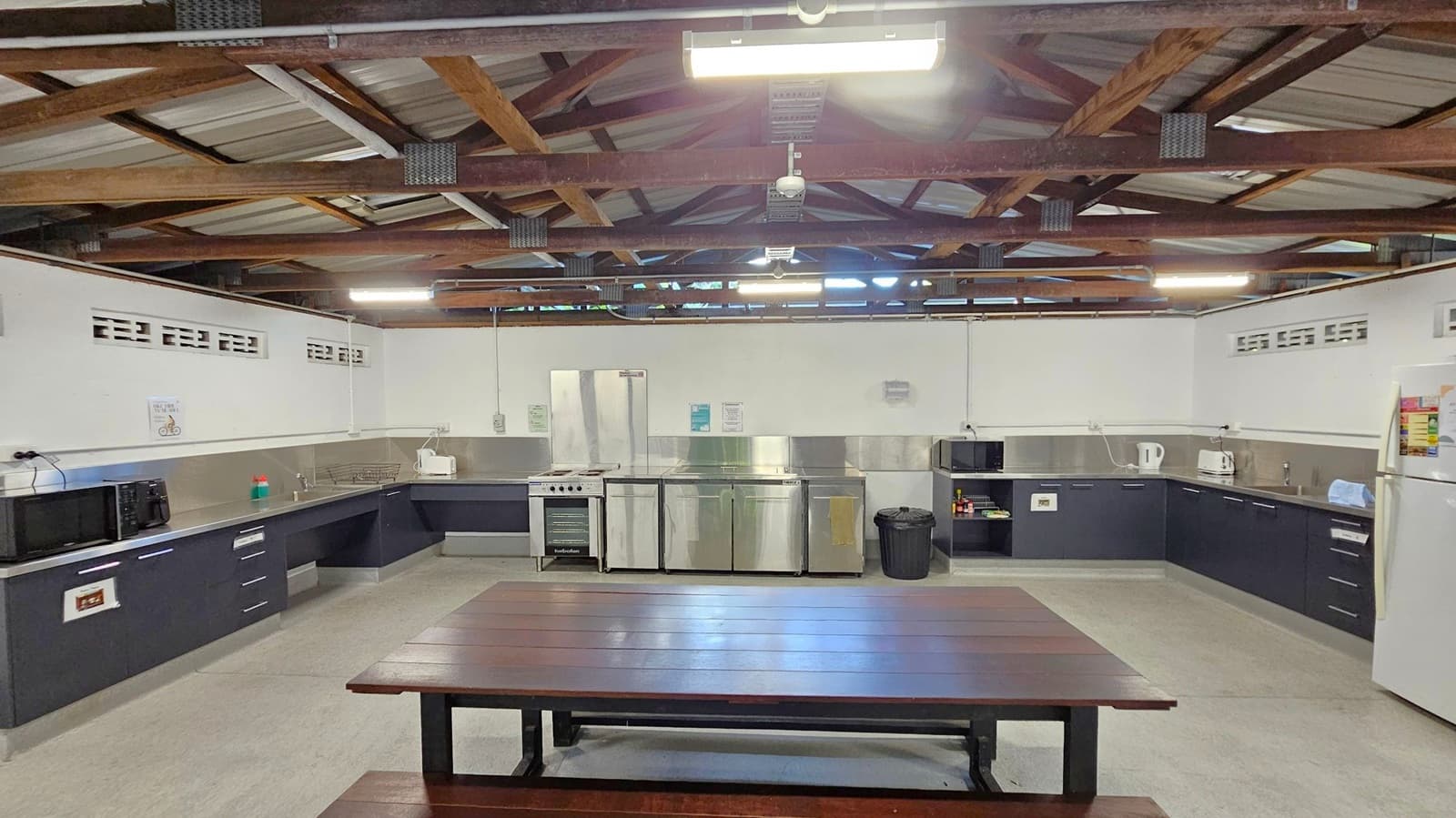 A spacious kitchen with a wooden picnic-style table in the foreground. The background features a U-shaped counter with black cabinets and stainless steel surfaces. Appliances include a microwave, toaster, kettle, and a large fridge on the right. The ceiling is open with exposed wooden beams and fluorescent lighting. The floor is light grey, and there are small ventilation blocks along the upper walls.