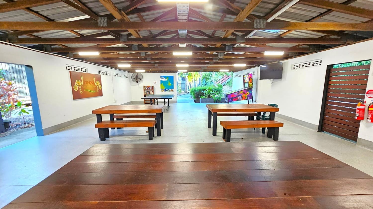 A spacious room with a wooden ceiling and exposed beams. In the foreground, there are wooden tables and benches. The left wall features a painting and a fan, while the right wall has a television and a wooden door with fire extinguishers nearby. The back of the room opens to an outdoor area with greenery and a pathway. The floor is concrete, and the walls are white with decorative ventilation blocks near the ceiling.