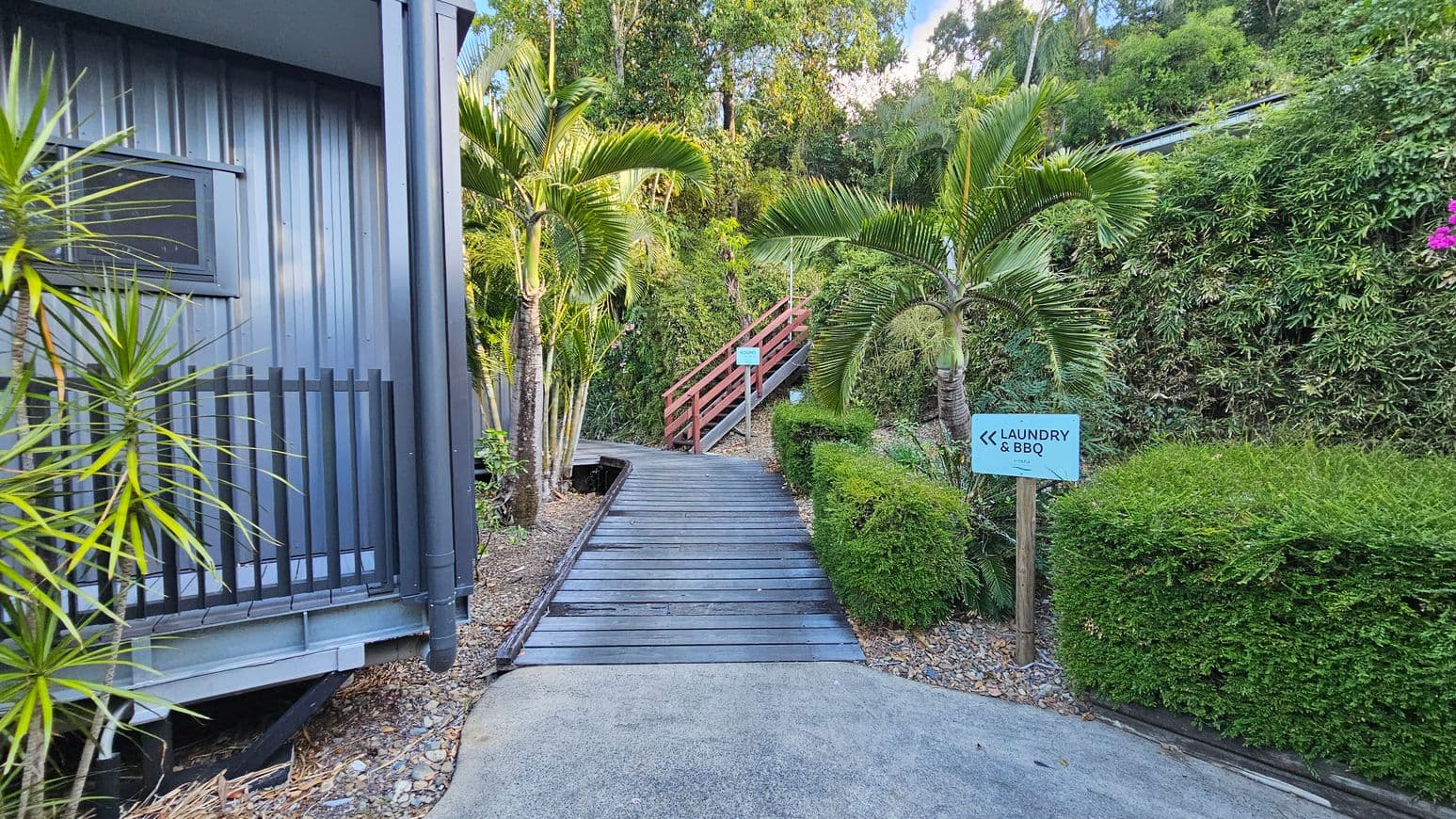 A pathway leads upwards, flanked by lush greenery and palm trees. On the left, a grey metal building with a small window and black railing is visible. On the right, a sign points towards "Laundry & BBQ" amidst trimmed hedges. The path transitions from concrete to wooden planks, leading to a red wooden railing. Dense foliage and trees form the background, creating a tropical atmosphere.