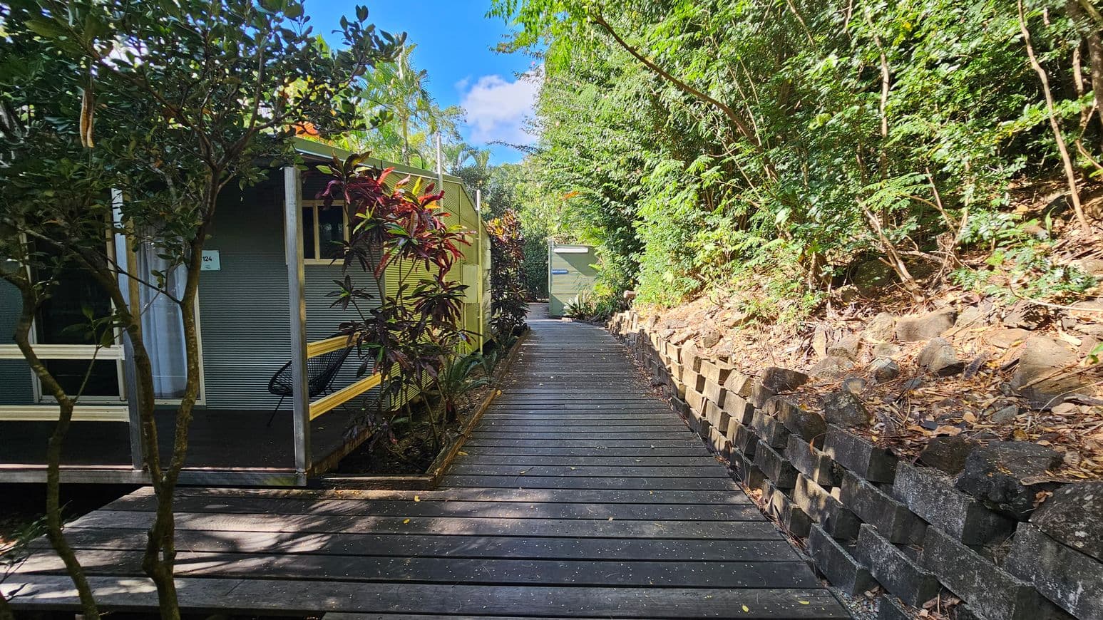 A narrow wooden walkway extends into the distance, flanked by lush greenery on both sides. On the left, a small building with corrugated metal walls and a porch is partially visible, surrounded by various plants. On the right, a stone retaining wall supports a slope covered with dense foliage. The path leads towards a small structure in the background, partially obscured by trees. The scene is well-lit, suggesting a sunny day.