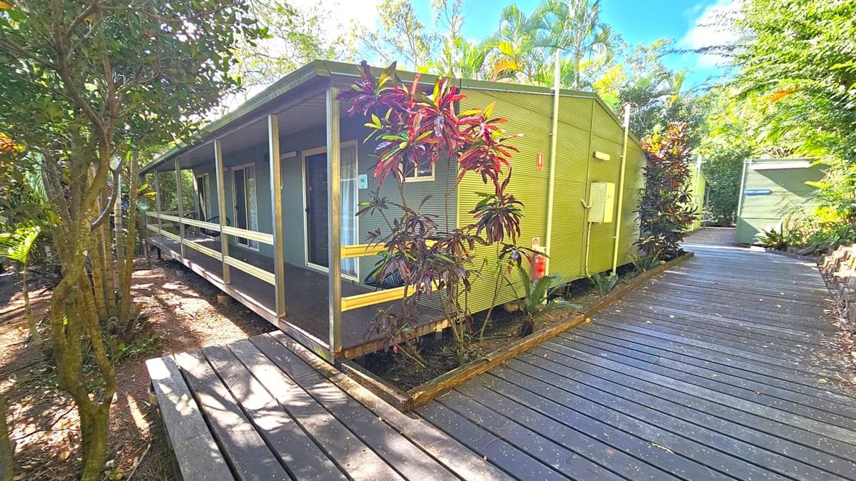 A wooden cabin with a corrugated exterior is on the left, featuring a small porch with railings. The cabin is surrounded by lush greenery, including trees and plants with red-tinted leaves. A wooden walkway runs alongside the cabin, leading to another structure in the background. The area is shaded by the surrounding foliage, and the ground is covered with mulch and leaves.