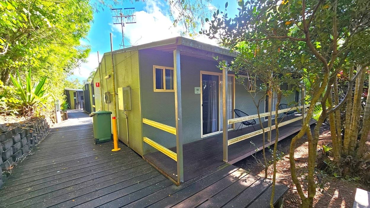 A small, corrugated metal cabin with a wooden deck and railing is on the right. It has a glass door and a small window. Two black chairs are visible on the deck. A green wheelie bin and a yellow bollard are near the cabin. A wooden walkway leads to another building in the background. Lush greenery surrounds the area, with trees and plants on both sides. The sky is partly visible above.