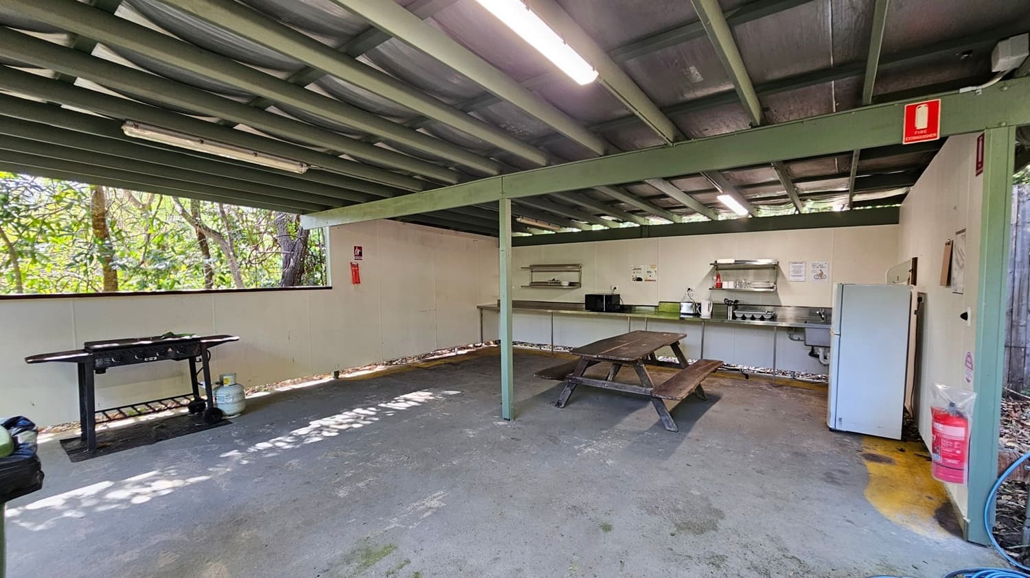 An outdoor kitchen area with a green metal roof and beams. On the right, there's a wooden picnic table and a white refrigerator. The back wall has a countertop with a microwave, kettle, and shelves. A fire extinguisher is mounted on the wall. On the left, a barbecue grill is placed near an open window with a view of trees. The floor is concrete, and the ceiling has fluorescent lights.