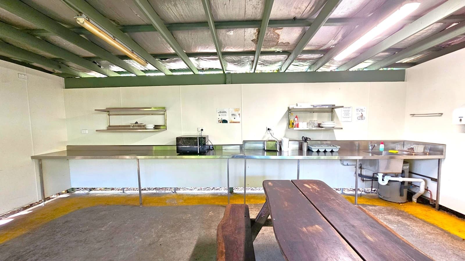 A simple kitchen area with a long stainless steel counter along the back wall. On the left, there are shelves with dishes and a toaster oven. In the centre, a sink with cleaning supplies and a dish rack. The ceiling has exposed beams and fluorescent lights. The floor is a mix of concrete and a worn carpet. In the foreground, a wooden picnic table is visible.