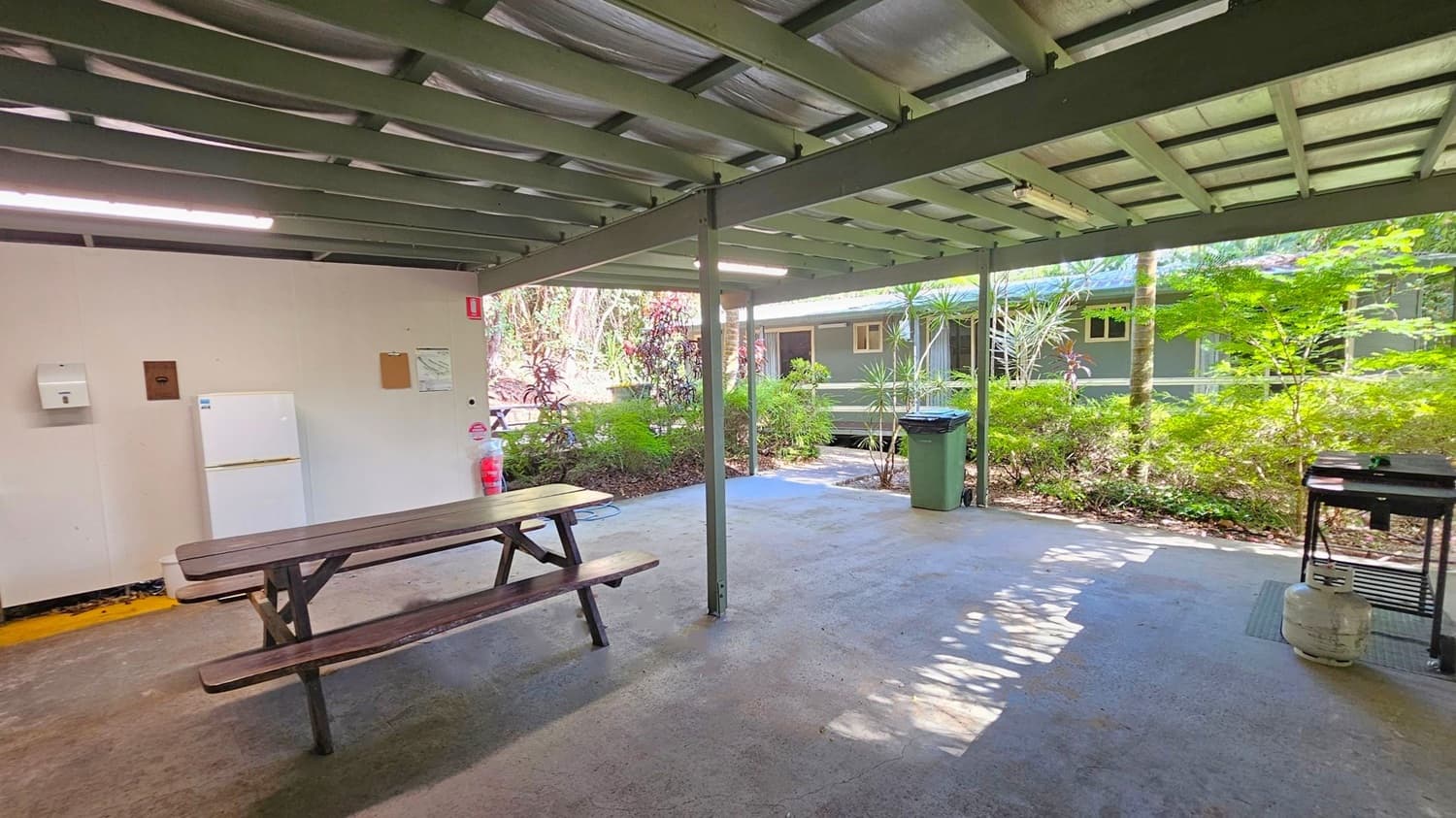 A covered outdoor area with a green metal roof and beams. On the left, there's a wooden picnic table and a white refrigerator against a beige wall. A fire extinguisher is nearby. In the middle, the floor is concrete with scattered leaves. On the right, a green rubbish bin and a barbecue grill with a gas cylinder are visible. The background features lush greenery and a building with windows.