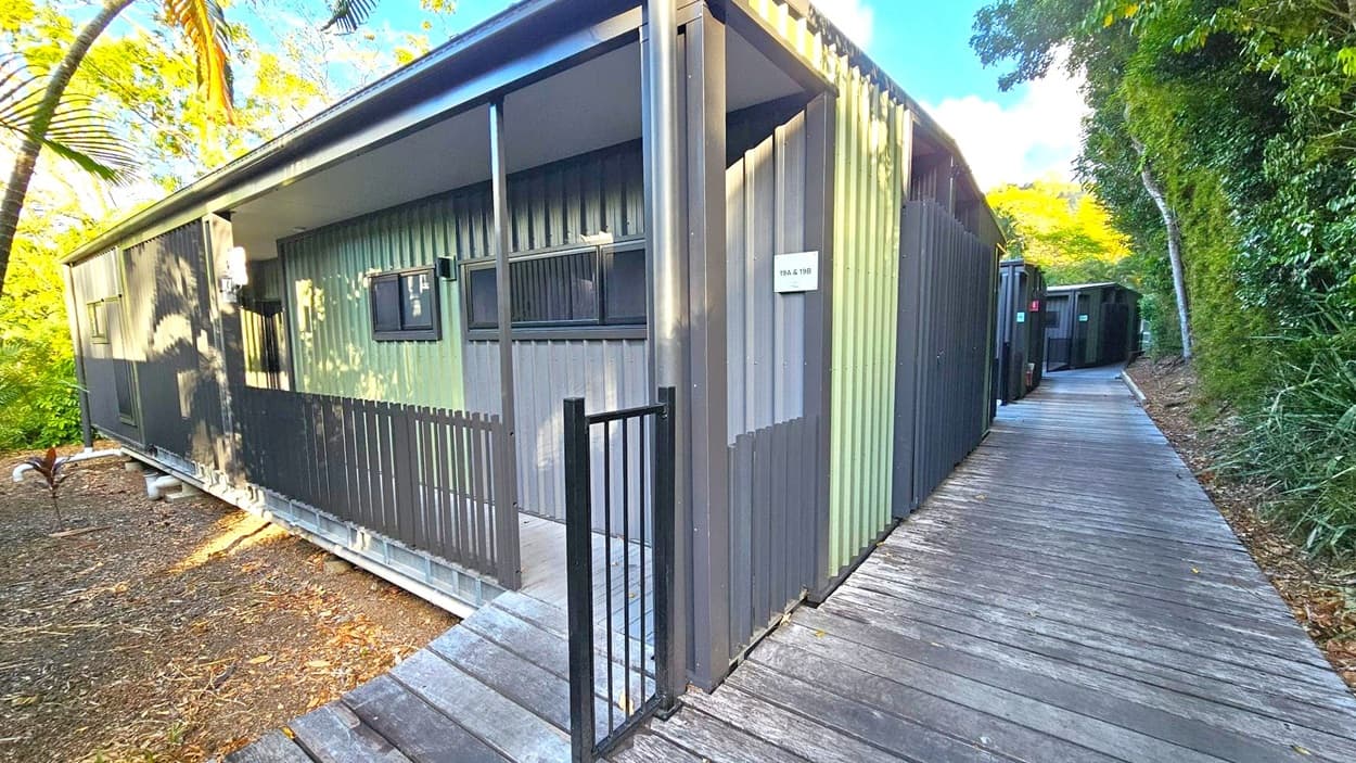 A modern building with corrugated metal walls in shades of grey and green is on the left. It features small rectangular windows and a black metal gate leading to a covered entrance. A wooden walkway runs alongside the building, curving into the distance on the right. The area is surrounded by lush greenery, including trees and bushes, with sunlight filtering through the leaves. The ground is covered with mulch and small plants.