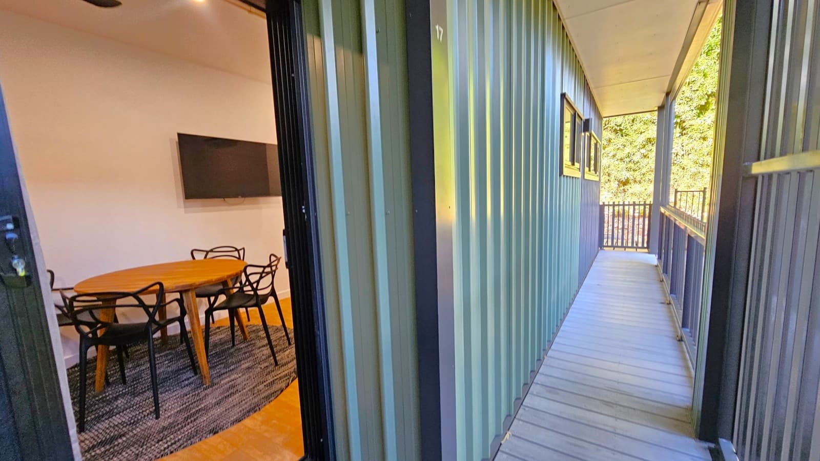 A narrow corridor with a wooden floor and metal walls on the right, leading to a railing at the end. On the left, an open door reveals a room with a round wooden table surrounded by black chairs on a dark rug. A flat-screen TV is mounted on the wall inside. The ceiling is light-coloured, and there are windows along the corridor wall.