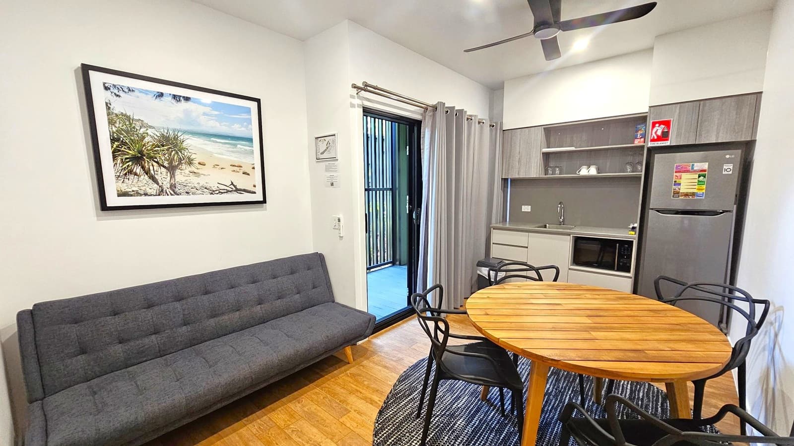 A compact living space with a grey sofa on the left, above which hangs a framed beach photograph. In the centre, a round wooden table with black chairs sits on a patterned rug. To the right, a kitchenette features a sink, microwave, and a fridge with a fire exit sign above. A ceiling fan is visible above. A sliding door with grey curtains leads to an outdoor area. The floor is wooden, and the ceiling is white.