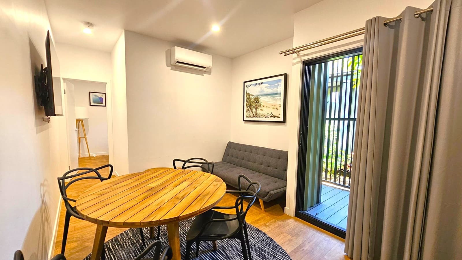 A room with wooden flooring and a light-coloured ceiling. On the left, a wall-mounted TV is visible. In the centre, a round wooden table with black chairs sits on a patterned rug. A grey sofa is against the back wall, beneath a framed beach scene. An air conditioning unit is above the sofa. On the right, a sliding glass door with vertical bars leads outside, partially covered by grey curtains. A hallway with a picture and a lamp is visible in the background.