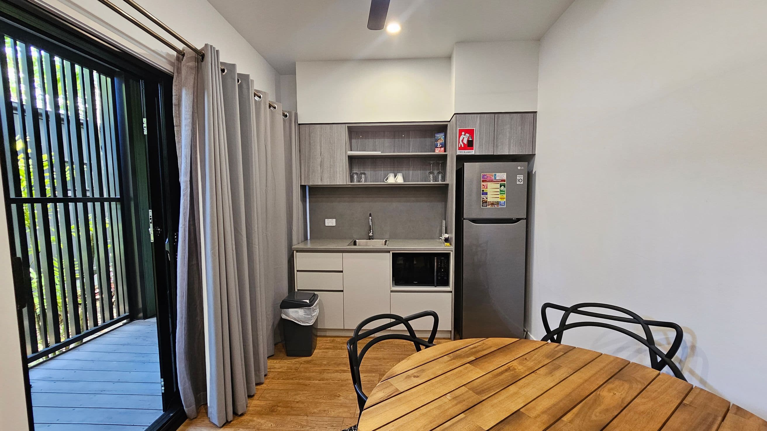 A compact kitchen area with a wooden floor. On the left, there is a sliding glass door with vertical black bars and beige curtains, leading to an outdoor area with visible greenery. In the centre, a kitchenette features a sink, a microwave, and open shelves with glasses and a few items. A small fridge is on the right, with a fire extinguisher sign above. In the foreground, a round wooden table with black metal chairs. The ceiling has a light and a fan.