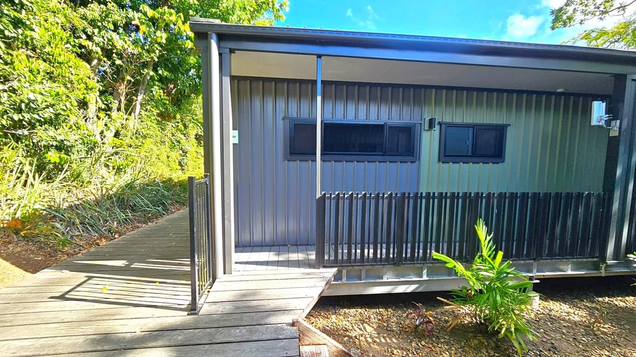 A small, modern cabin with a metal exterior and a covered porch. The porch has a black railing and a wooden walkway leading to it. Two rectangular windows are visible on the cabin's front. On the left, dense green foliage and trees provide a natural backdrop. The ground is covered with gravel and a few small plants. The sky is not visible.