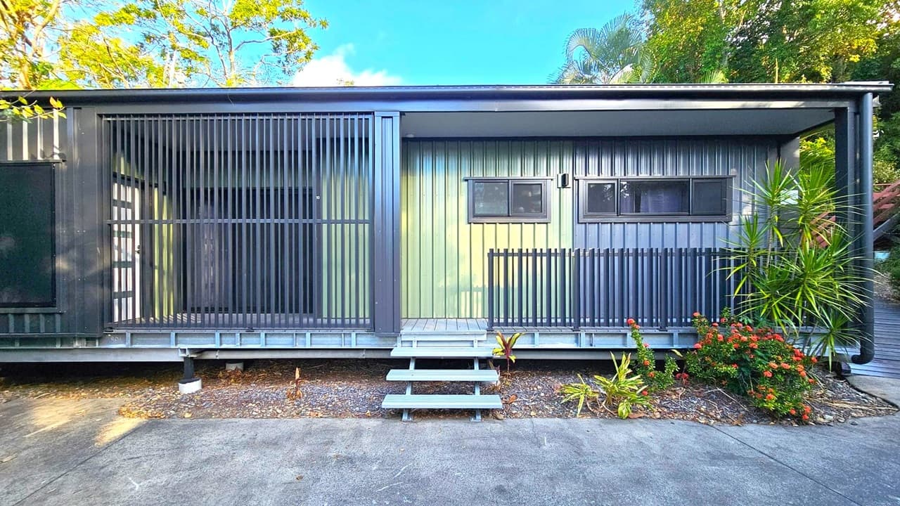 A modern building with a metal exterior featuring vertical slats on the left side. In the centre, there are two rectangular windows with a small awning above. A set of grey steps leads up to the entrance. On the right, there are plants, including a tall, spiky one and a bush with red flowers. The ground is covered with gravel and a concrete path. Trees and foliage are visible in the background.