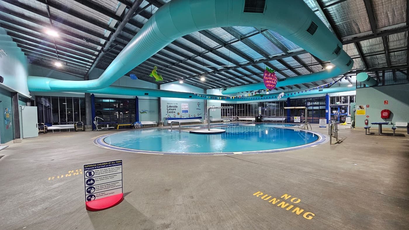A glass entrance to a swimming pool area with blue-framed doors. On the left, a board displays photos and names of swim teachers at the Lakeside Leisure Centre. Below it, a white wire basket with a red sale sign. On the right, a vertical banner promoting "Float to Survive with GOswim" stands next to the door. Through the glass, a pool with turquoise pipes and aquatic-themed decorations is visible. The floor is grey, and the ceiling has exposed beams and lights.