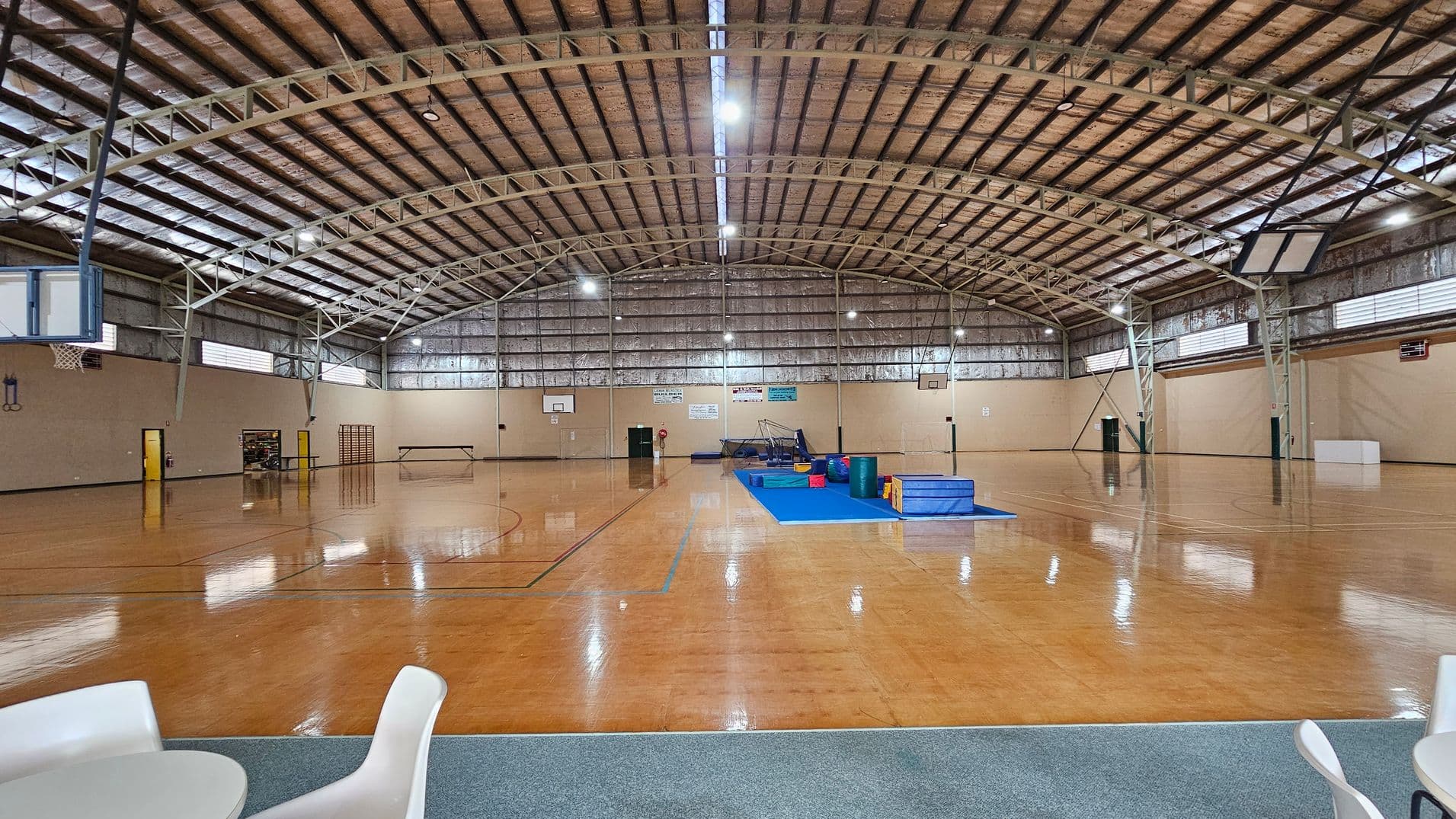 A large indoor sports hall with a high, arched metal ceiling. The floor is polished wood, marked for basketball. In the centre, there's a blue mat with gymnastic equipment. On the left, a basketball hoop is visible, and on the right, another hoop and a white box. The walls are lined with windows near the ceiling. In the foreground, there are white chairs and a table on a carpeted area.