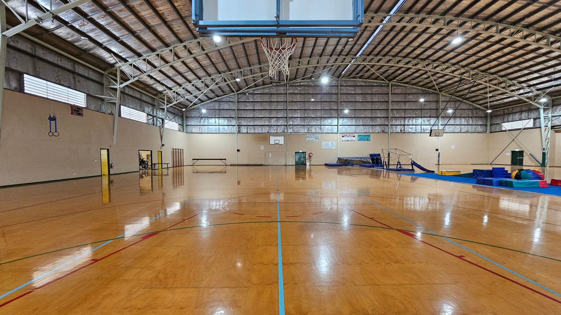 A spacious indoor gymnasium with a polished wooden floor and a high, arched metal ceiling. In the centre, a basketball hoop is suspended. The left side has a wall with small windows and a few doors, along with a bench. On the right, there are gymnastic mats and equipment, including a balance beam and vaulting horse. The back wall features a basketball hoop and several signs. The lighting is bright, with multiple ceiling lights.