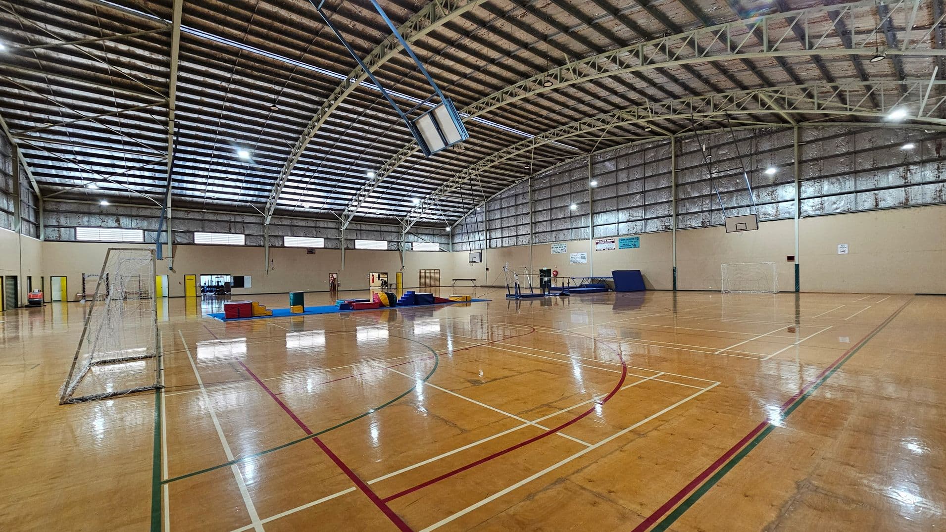 A large indoor sports hall with a polished wooden floor marked for various sports. On the left, there's a football goal. In the centre, gymnastic equipment like mats and vaulting horses are arranged. The right side has a basketball hoop attached to the wall. The ceiling is high with metal beams and lights, and the walls are lined with windows and ventilation panels.