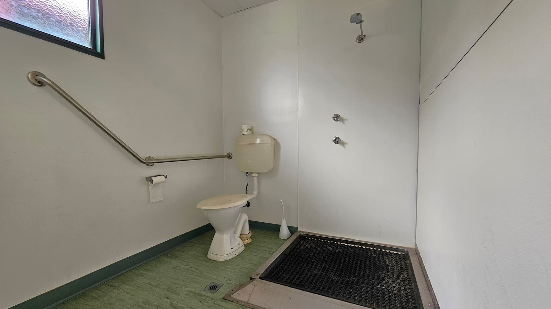 A bathroom with light green walls and a green floor. On the left, there's a toilet with a beige cistern, a toilet brush, and a roll of toilet paper on a holder. A metal grab bar runs along the wall. On the right, a shower area with a black rubber mat and three wall-mounted shower controls. A small window with frosted glass is visible in the upper left corner. The ceiling is plain and light-coloured.