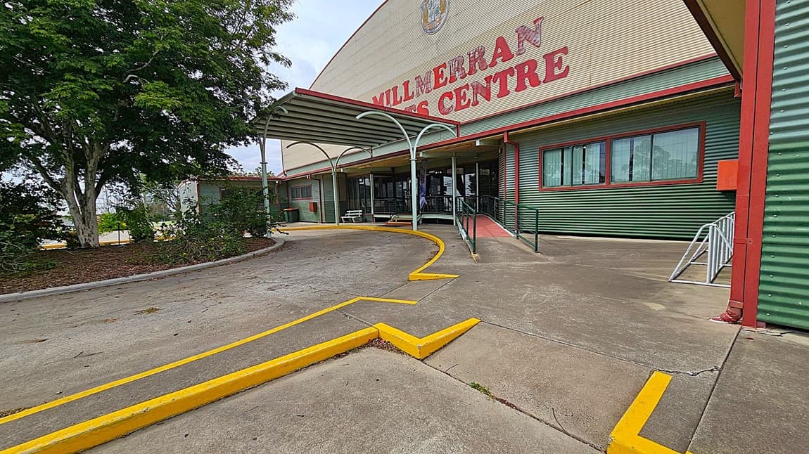 A large building with a corrugated metal exterior painted green and beige. The words "Millmerran Centre" are prominently displayed in red on the upper part of the building. A covered entrance with a metal awning leads to glass doors. A concrete path with yellow-painted edges curves towards the entrance. On the left, there is a large tree with dense foliage. The ground is a mix of concrete and asphalt, with some scattered leaves.