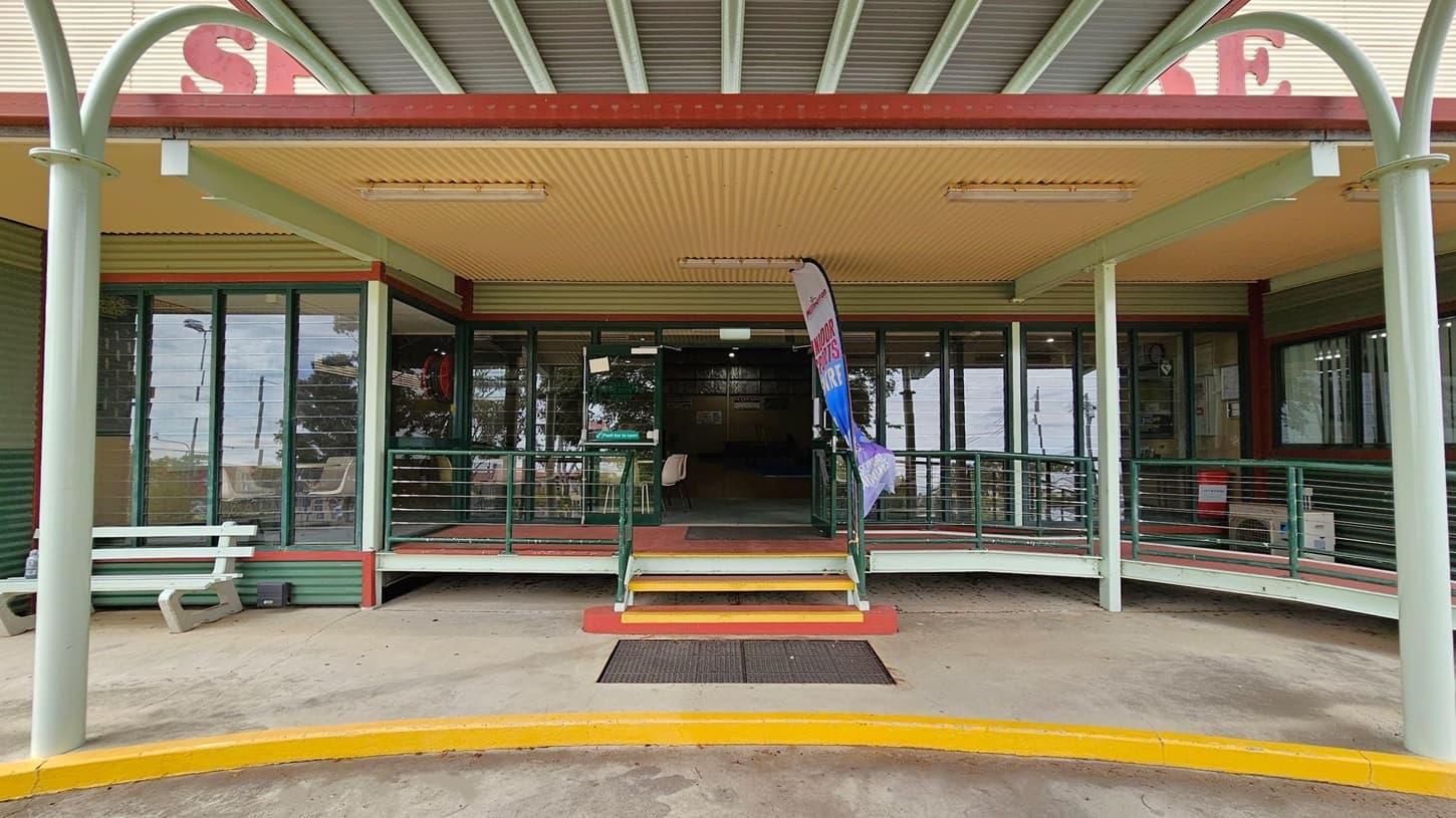 A building entrance with a covered porch supported by light green pillars. The porch has a corrugated metal ceiling and fluorescent lights. A set of steps with yellow edges leads up to the entrance, flanked by a metal railing. A flag banner stands to the right of the steps. Large glass windows with louvres are on either side. A white bench is on the left, and the ground is concrete with a yellow-painted kerb.