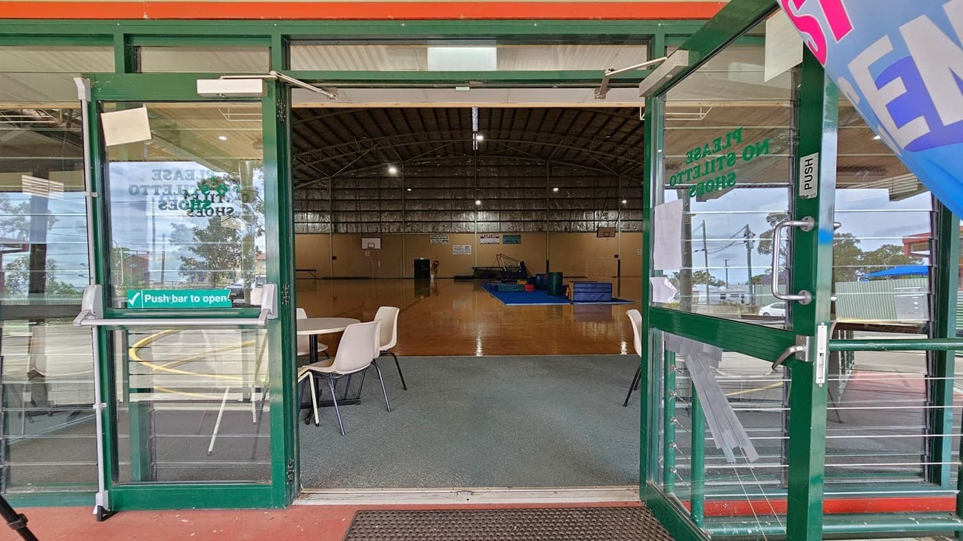 Green double doors with glass panels are open, leading into a large indoor sports hall. The floor is polished wood, and the ceiling is arched with metal beams. Inside, there are gym mats and equipment on the right. In the foreground, a round table with white chairs is visible. Signs on the doors read "Please No Stiletto Shoes" and "Push bar to open." Outside, a blue and pink flag is partially visible on the right.