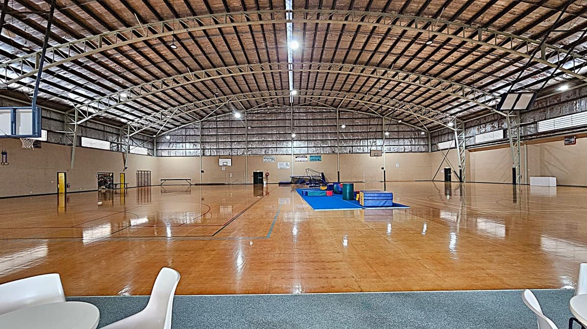 A large indoor sports hall with a high, arched metal ceiling. The floor is polished wood, marked with sports lines. In the centre, there's a blue mat with gym equipment. On the left, a basketball hoop and some gymnastic rings are visible. On the right, another basketball hoop and a white box. The walls are beige with high windows. In the foreground, a small area with a grey carpet and white chairs and tables.