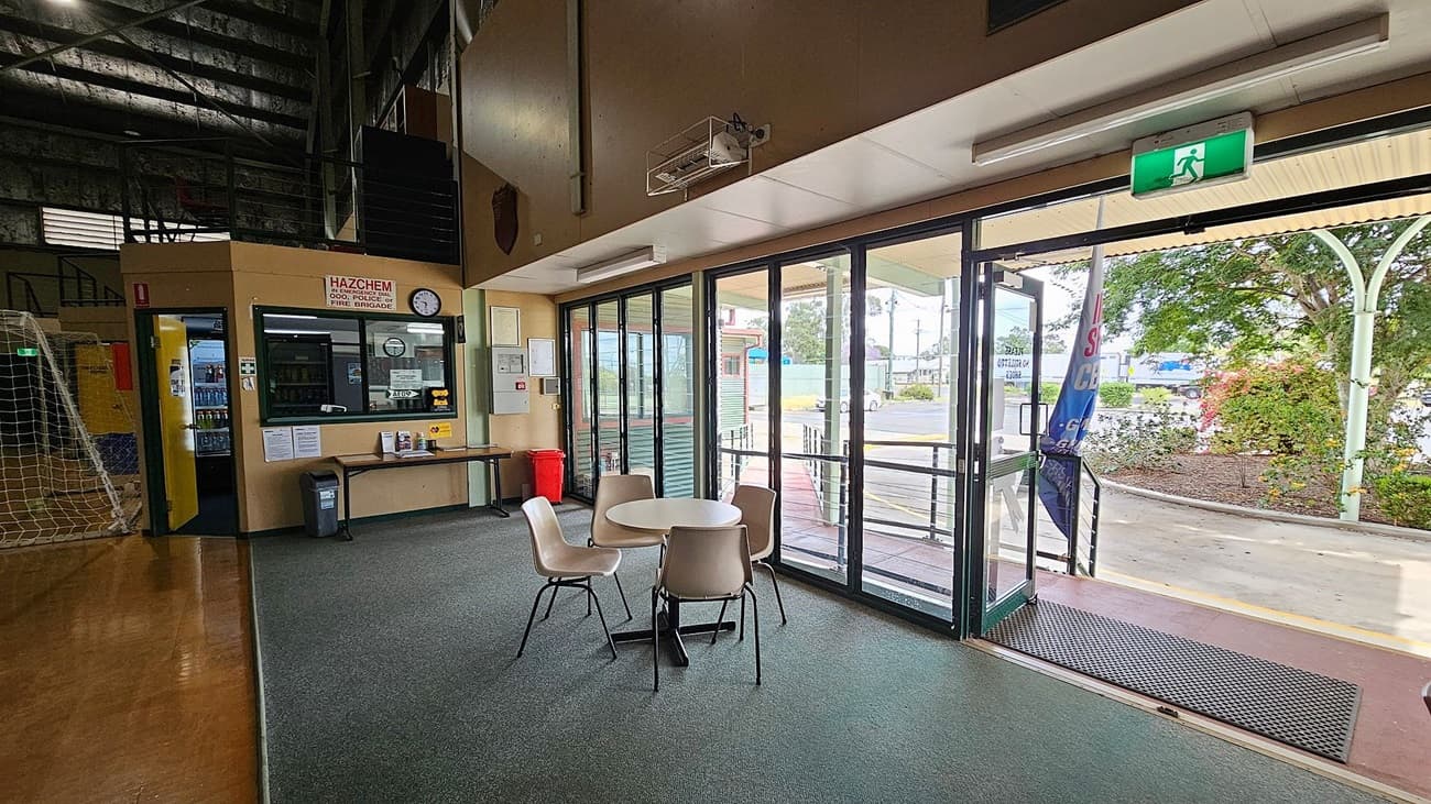 Inside a building with a high ceiling, a small seating area with a round table and four chairs is on a grey carpeted floor. To the left, there's a counter with a window, a clock, and a "HAZCHEM" sign above. A vending machine is visible nearby. Large glass doors on the right lead outside, where a garden with bushes and a tree is visible. An exit sign is above the doors, and a rubber mat is on the floor at the entrance.