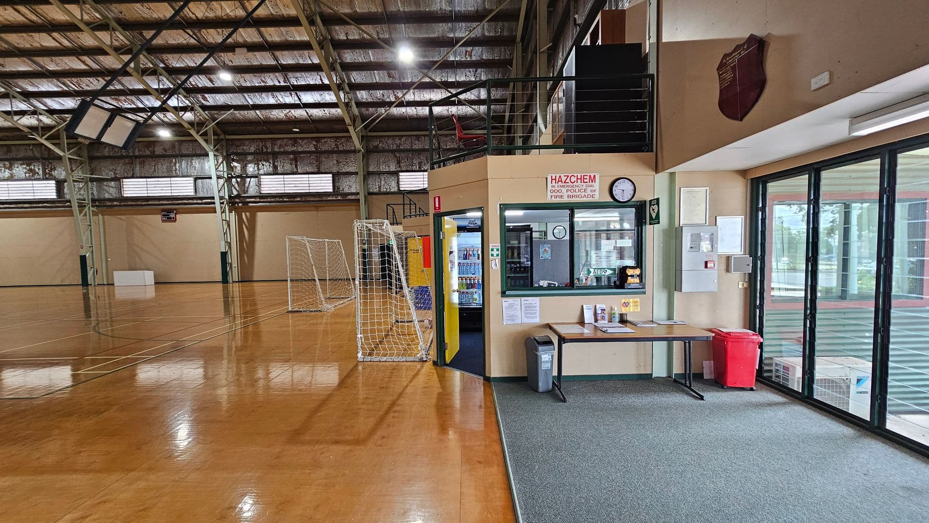 A sports hall with a polished wooden floor and high metal ceiling. On the left, there are two white goal nets and a basketball hoop. In the centre, a small office area with a window, clock, and emergency sign. A table with papers and a red bin are nearby. On the right, large glass doors with a view of the outside. The ceiling has exposed beams and lights, and the floor is shiny and reflective.