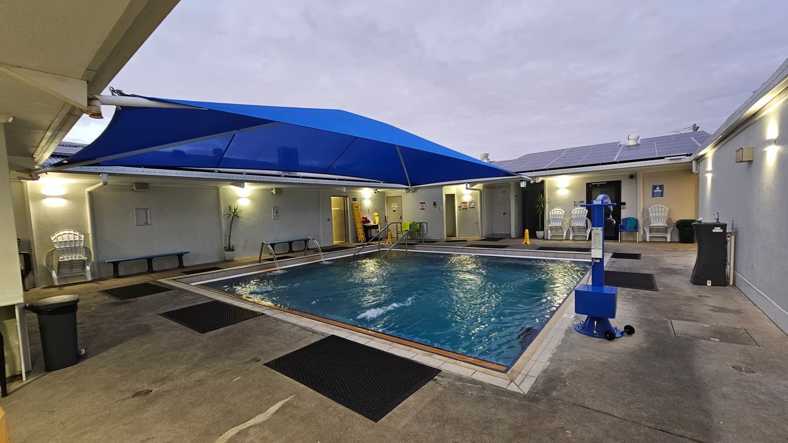 A sliding glass door is partially open, revealing a pool area with a small pool and blue equipment. The left side of the door is frosted with a pattern. On the left wall, there is a historical display with images and text. The right wall has a similar display labelled "TREATMENT ROOMS." The floor has black mats, and a yellow caution sign is visible near the pool. The ceiling is white with lights.