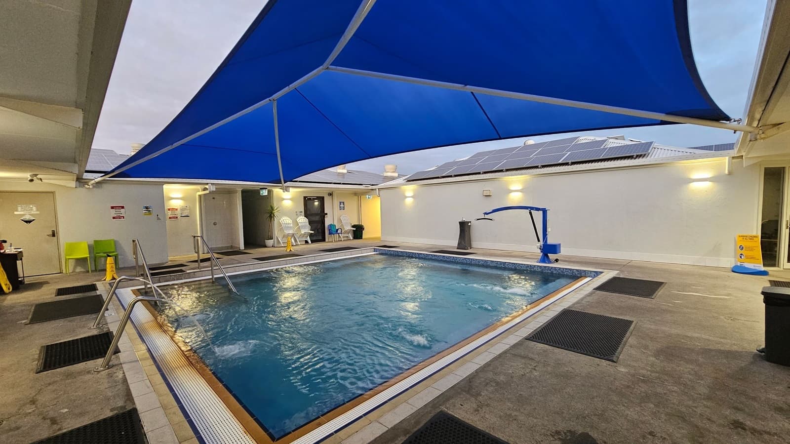 An indoor swimming pool area with a blue shade sail overhead. The pool has metal handrails and is surrounded by a tiled deck with black mats. On the left, there are green chairs and a door with signs. The back wall has white plastic chairs, a potted plant, and a door. On the right, a pool lift is visible. The floor is concrete, and the ceiling is partially visible with solar panels on the roof.