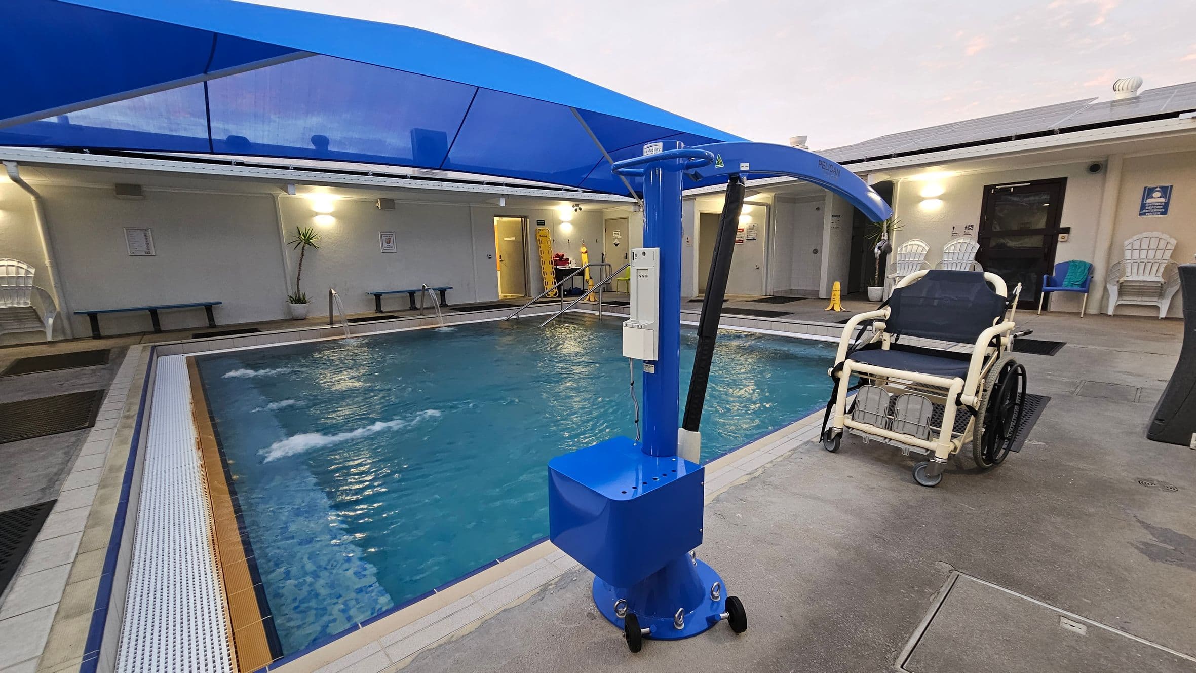 An indoor swimming pool with a blue hoist on the right, next to a wheelchair. The pool has a tiled edge and water jets creating ripples. On the left, there are benches and a potted plant against the wall. The background shows a building with doors, windows, and white chairs. A blue canopy covers the pool area. The floor is a mix of tiles and concrete.
