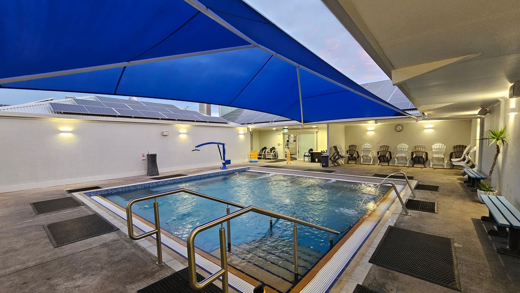 An indoor swimming pool with a blue canopy overhead. The pool has metal handrails and is surrounded by non-slip mats. On the right, there are several white and brown plastic chairs lined up against the wall, with a clock above them. A small plant is in the corner. The left side features a wall with solar panels on the roof. The floor is concrete, and the ceiling is partially covered by the canopy.