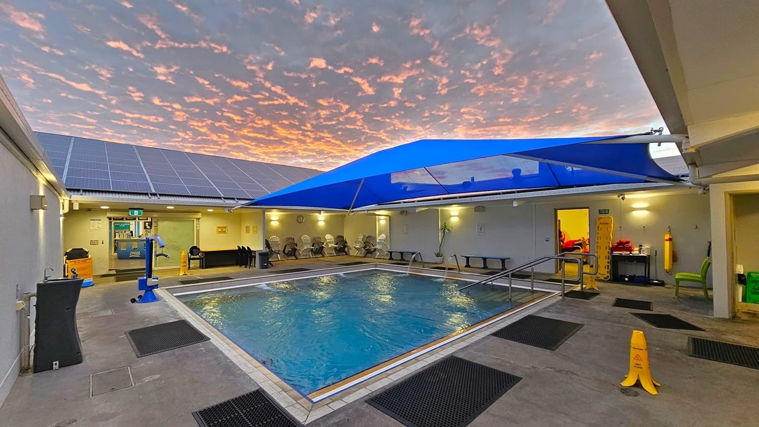 Indoor swimming pool area with a blue shade canopy above. The pool is surrounded by a tiled deck with black mats. On the left, there is a sink and a blue pool lift. The back wall has solar panels on the roof and a row of chairs under a lit overhang. On the right, an open door leads to a room with equipment. A yellow caution sign is on the floor near the pool. The ceiling is not visible.