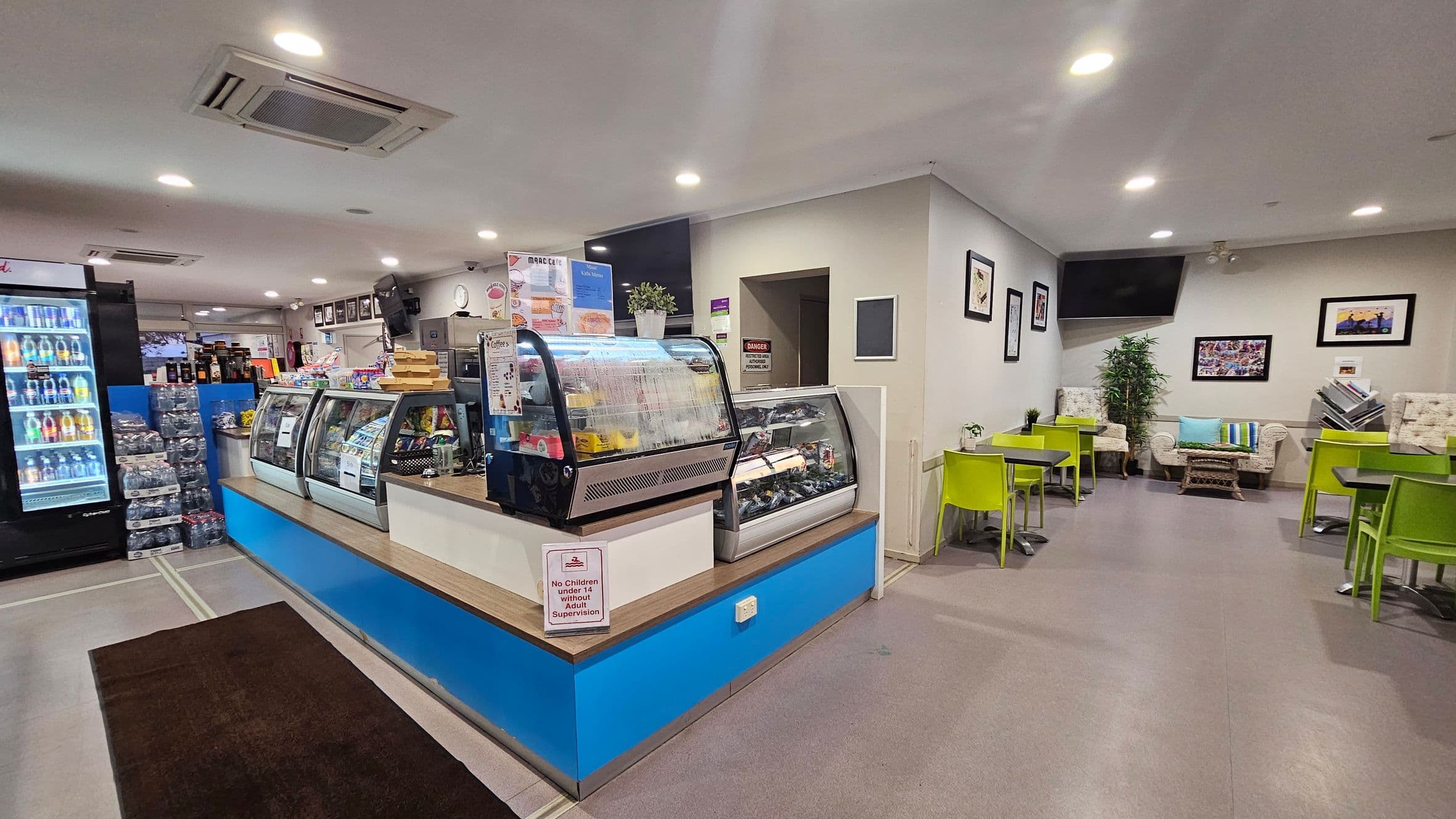 A spacious indoor area with a café counter in the centre, featuring a display of snacks and a sign about plastic bottles. To the right, there are lime green tables and chairs, along with a small seating area with sofas and a plant. On the left, there are turnstiles and a corridor leading to another section. The floor is tiled, and the ceiling has recessed lighting and air conditioning units. Walls are decorated with framed pictures.