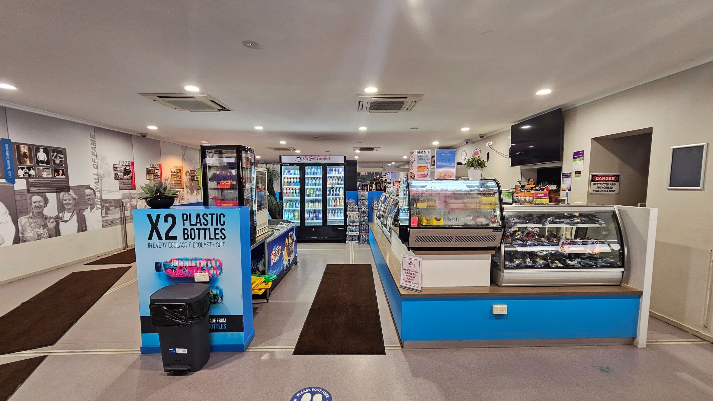 A convenience store interior with a grey ceiling and floor. On the left, a wall features historical photos and text. In the centre, there are refrigerated display units filled with drinks. A sign about plastic bottles is in front of a bin. On the right, a counter displays snacks and a menu board is visible. A "Danger" sign marks a restricted area. The store is well-lit with ceiling lights.