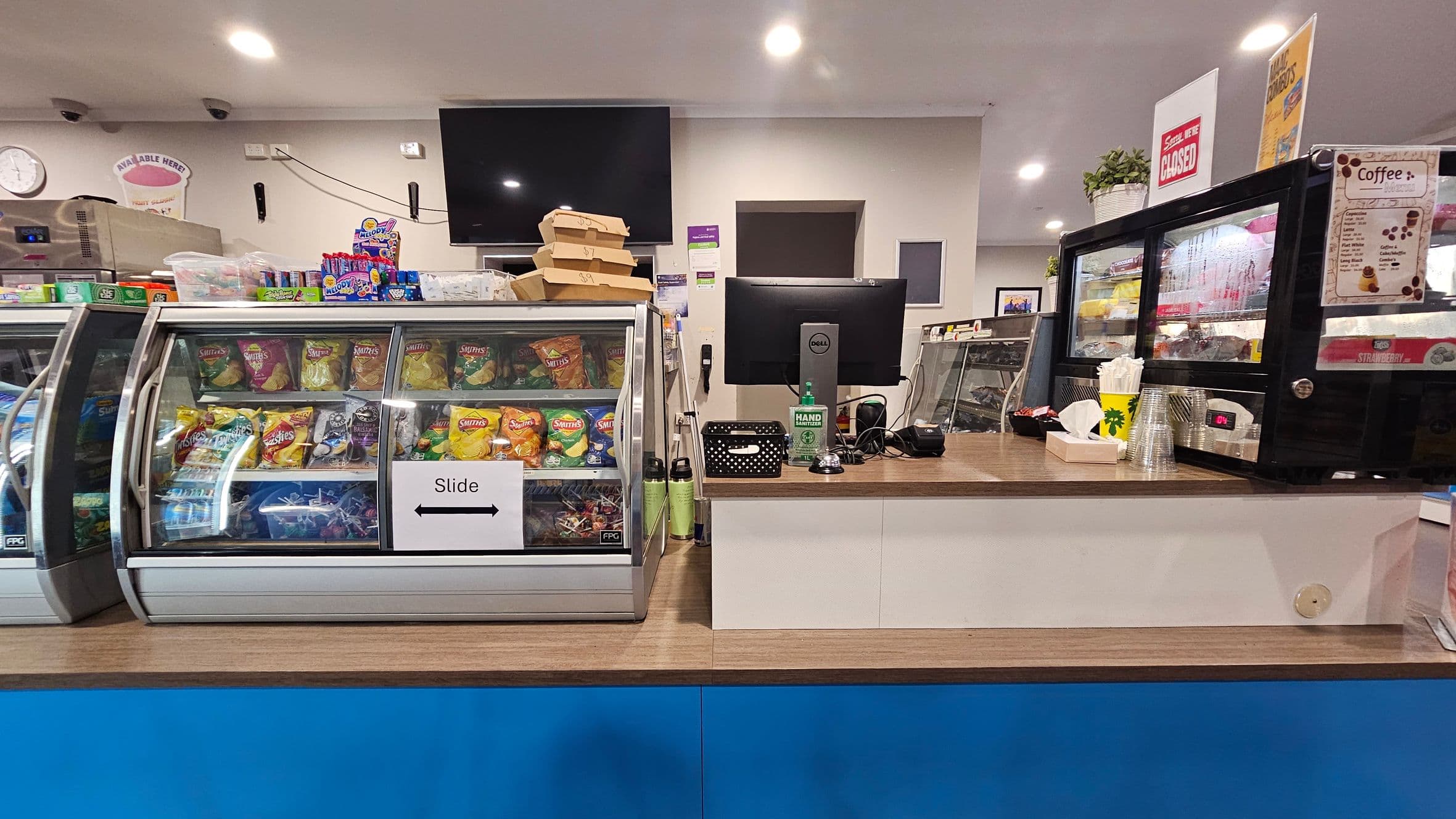 A shop counter with a glass display case on the left filled with various crisps and snacks. Above it, there are boxes and confectionery items. In the centre, a computer monitor sits on the counter with a bottle of hand sanitiser and a small basket. On the right, a black display case contains more snacks and a coffee menu is visible. The counter is wooden with a blue front, and the ceiling has recessed lighting. A "Sorry, We're Closed" sign is displayed above.