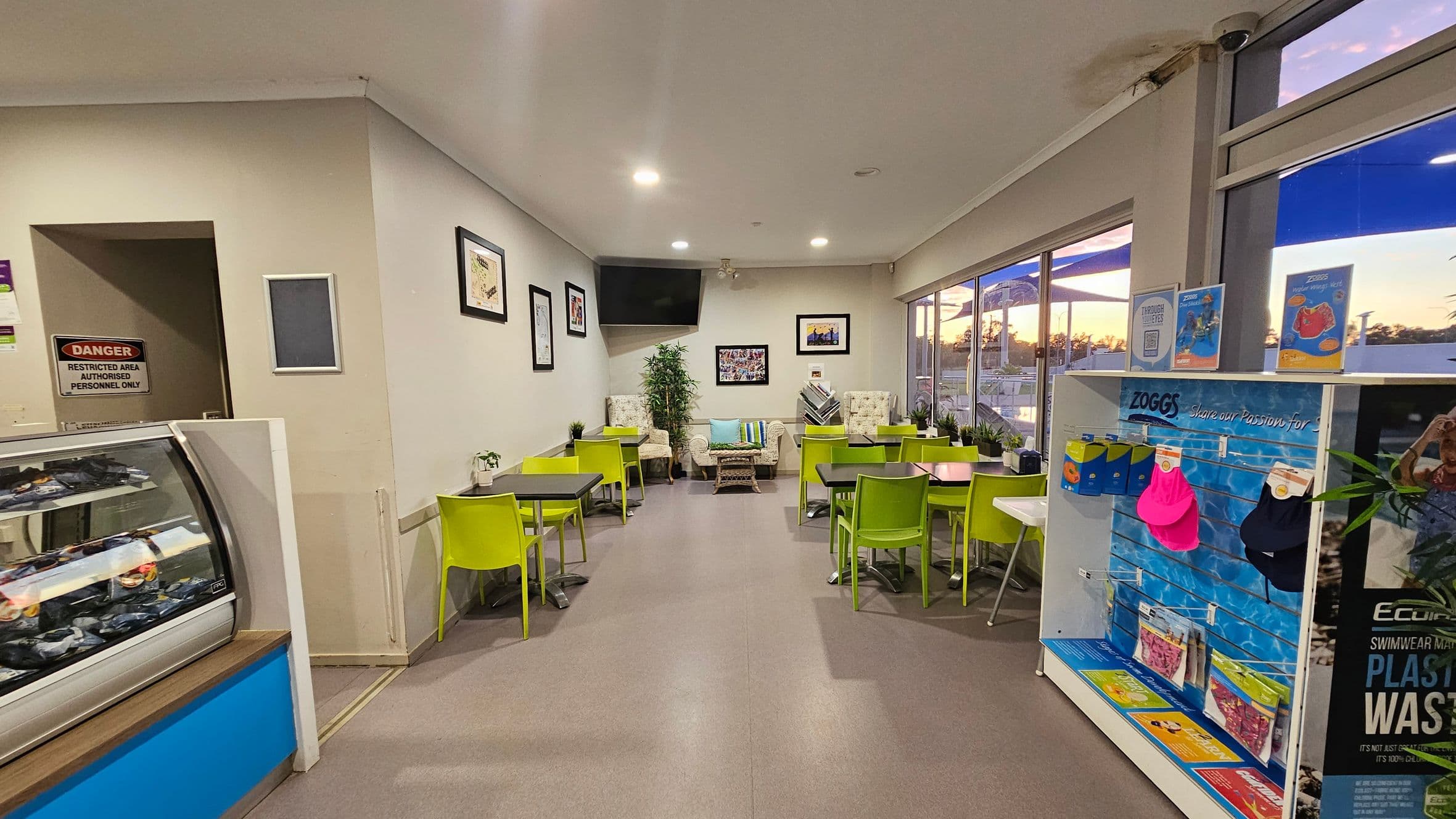 A small café with lime green chairs and black tables arranged neatly. On the left, there's a refrigerated display case and a door marked "Danger: Restricted Area." The back wall features framed pictures and a large TV. Two patterned armchairs and a small table with a plant are in the centre. On the right, a display shelf holds swimming gear. Large windows on the right show an outdoor view with a sunset. The floor is grey, and the ceiling is white with recessed lighting.