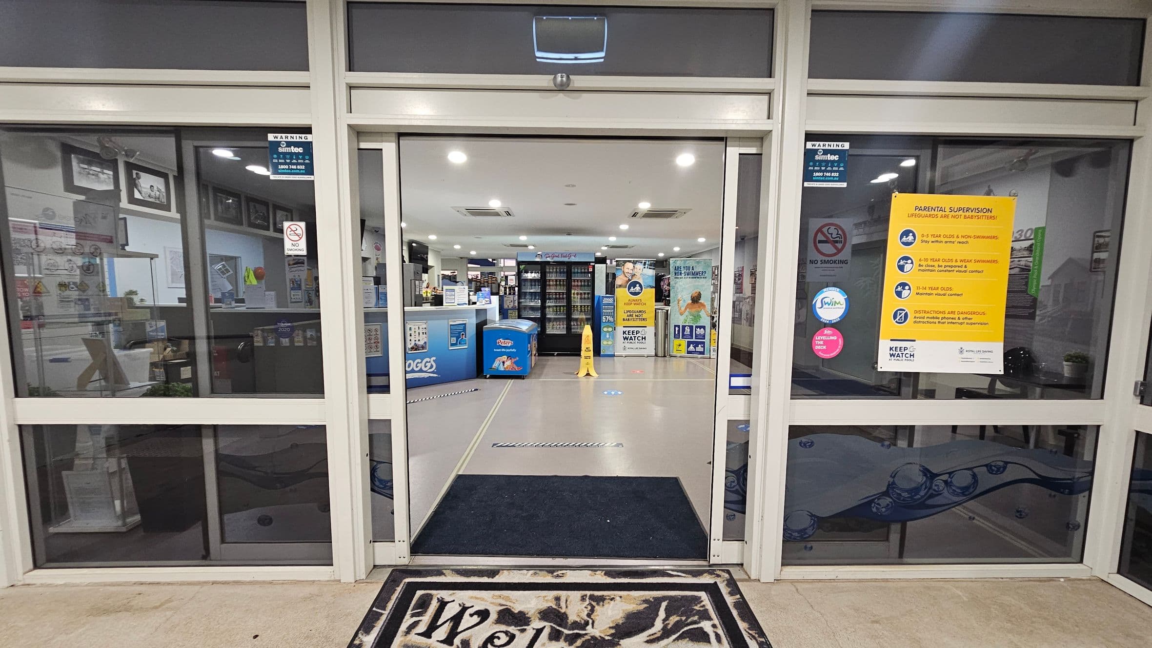 Glass entrance doors with a "Welcome" mat in front. On the left, there are posters and a "No Smoking" sign. On the right, a yellow sign about parental supervision. Inside, a blue counter with "Zoggs" branding is visible, along with a freezer and shelves stocked with items. The floor is tiled, and the ceiling has recessed lighting. A yellow caution sign stands in the centre.