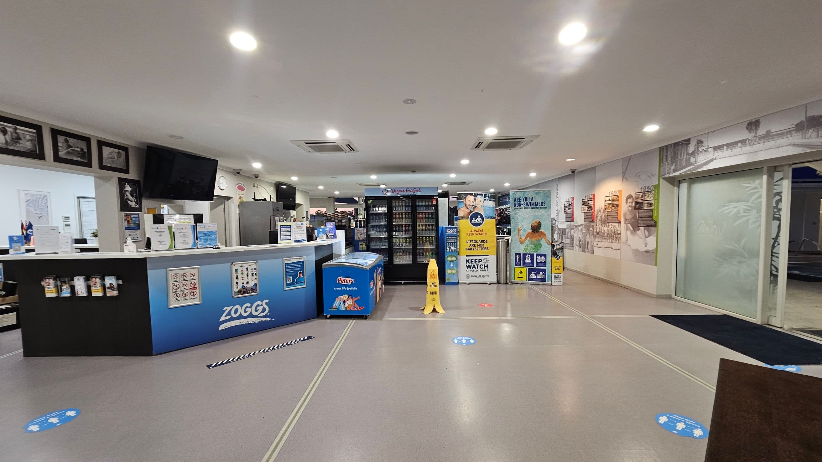 A reception area with a counter on the left displaying brochures and signs, branded with "Zoggs". Behind the counter are framed photos and a wall-mounted TV. In the centre, there's a freezer and a drinks fridge. On the right, a wall features historical images and a glass door. The floor is marked with social distancing stickers, and a yellow caution sign is present. The ceiling has recessed lighting.